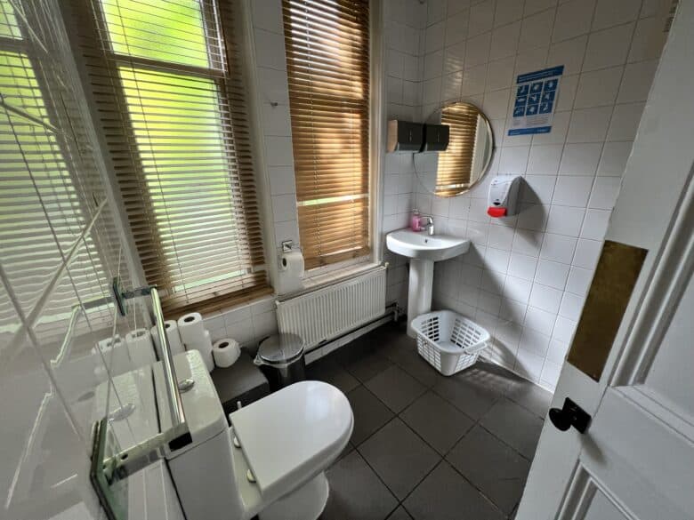 A small bathroom with white tiled walls, a window with wooden blinds, a toilet, a white pedestal sink with a round mirror above it, a soap dispenser, a wall-mounted hand drier, a radiator, and a laundry basket on the floor.