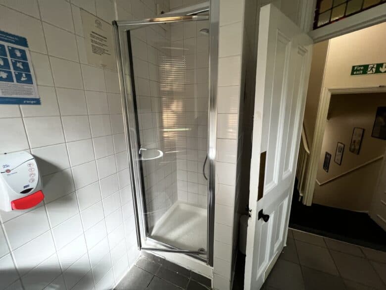 A small, enclosed shower with a glass door is situated in a tiled bathroom. An informational sign and a soap dispenser are mounted on the wall to the left of the shower. An open door reveals a hallway with a "Fire exit" sign.