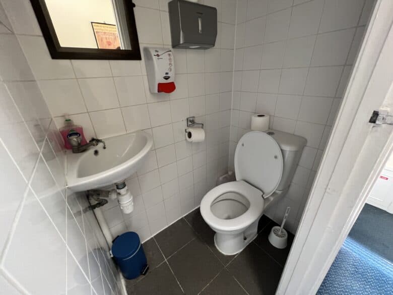 A small, clean bathroom with white tiled walls and gray tile floor. Features a toilet with the lid open, wall-mounted soap dispenser, hand sanitizer, sink with liquid soap, paper towel dispenser, toilet paper holder, toilet brush, and a blue trash bin.