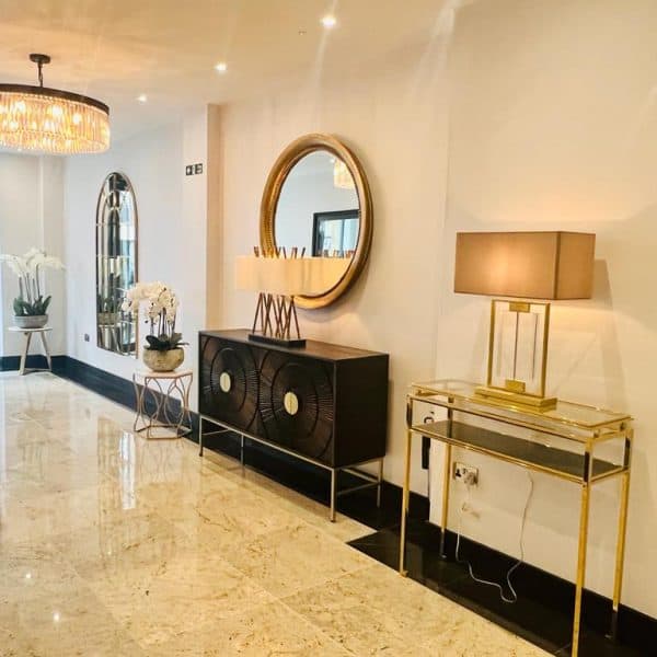 A modern, well-lit lobby in one of the finest hotels in London, boasting a polished marble floor. The space features a chandelier, large round mirrors on a white wall, decorative plants, and stylish furniture including a black sideboard and a glass-top table with a lamp. There are windows and a green exit sign.