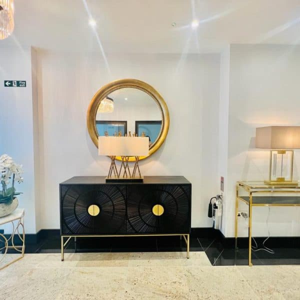 Modern interior featuring a black console table with gold accents, adorned with a lamp and a large round mirror above it. To the left, there's a small round table with white orchids. To the right, a glass table with another lamp. Bright lighting enhances the decor, reminiscent of luxurious hotels in London.