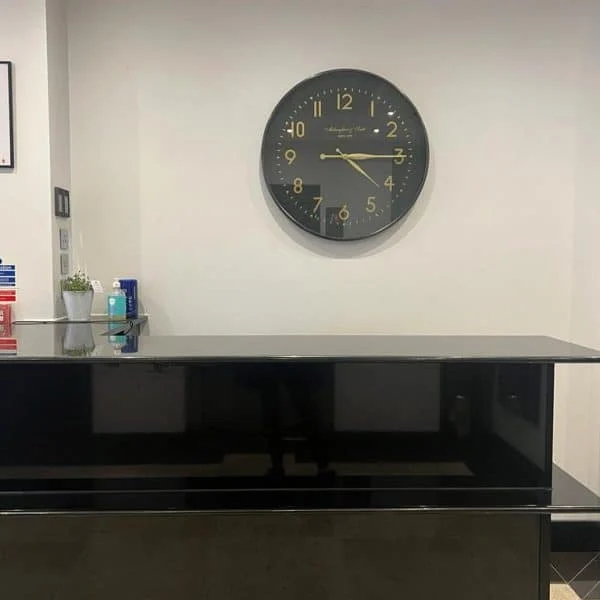 A reception desk with a glossy black counter is in front of a white wall, typical of hotels in London. A large black clock with gold numbers hangs on the wall behind the desk. The counter has a hand sanitizer bottle, a blue cup, and a small potted plant. Various notices and equipment are seen nearby.