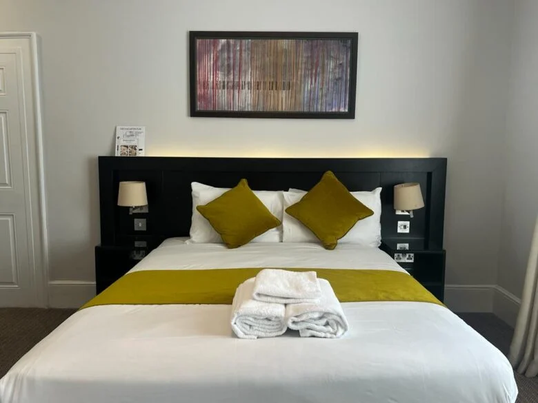 A modern hotel room featuring a neatly made bed with white linens, green accent pillows, and a matching green bed runner. Two rolled white towels are placed on the bed. The headboard includes built-in reading lamps, and a framed artwork hangs above the bed.