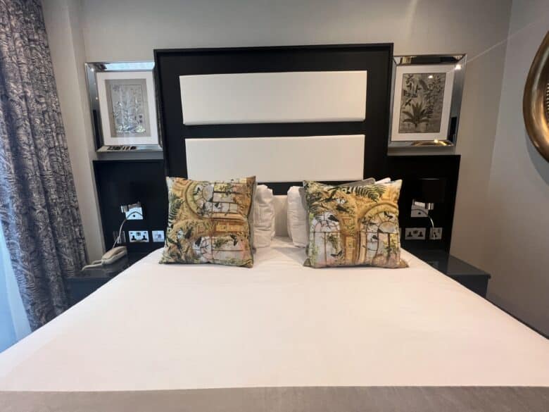 A hotel room features a neatly made bed with white linens and two decorative pillows adorned with nature-themed designs. The headboard has a modern black and white design, and framed artwork is displayed on the wall. Nightstands flank the bed, and a patterned curtain is partially visible.