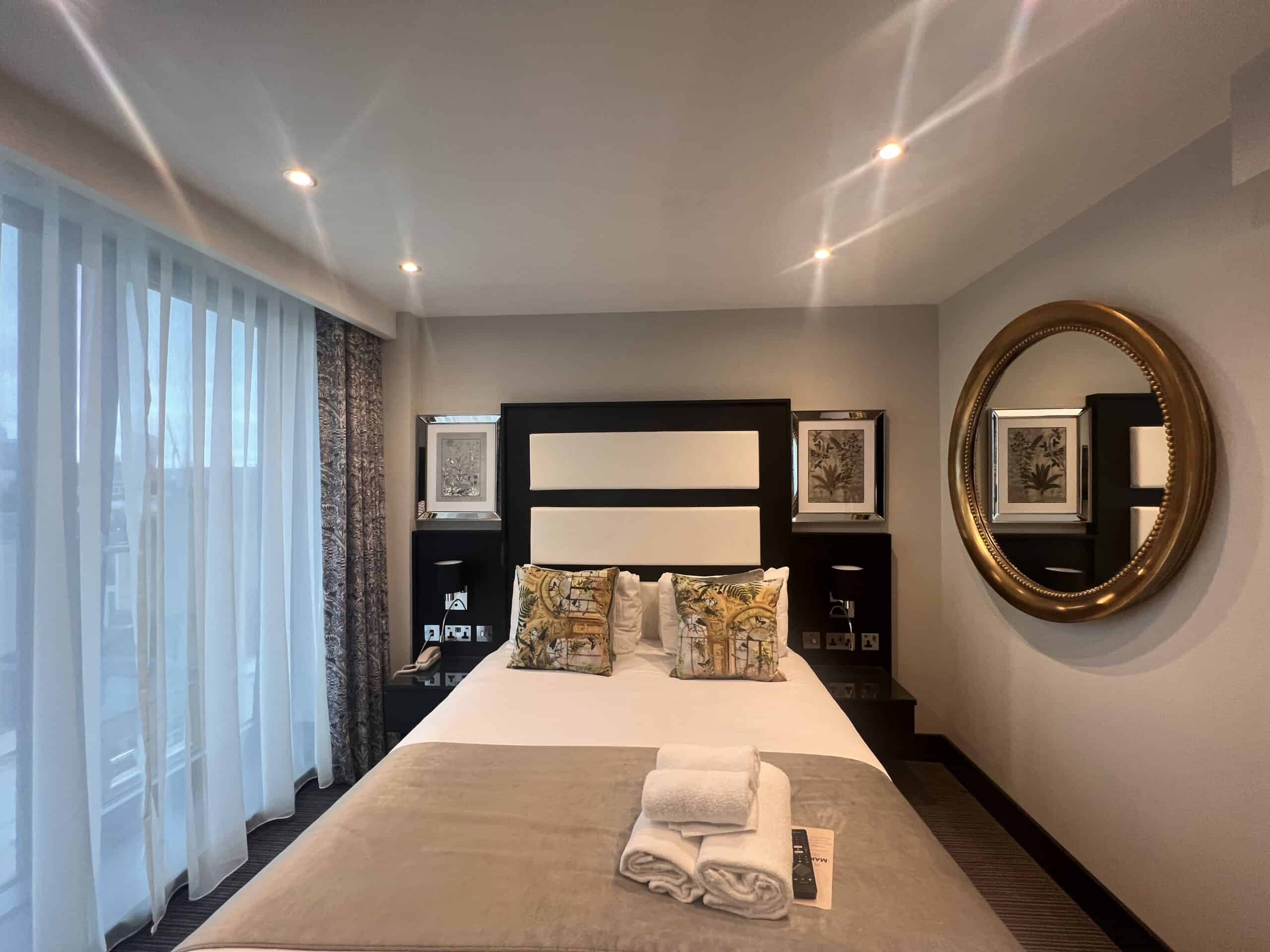 A modern hotel room featuring a neatly made double bed with patterned pillows, a folded blanket, and fresh towels on top. The room has a large round mirror on the right wall, artwork above the bed, and a window with sheer white curtains on the left.