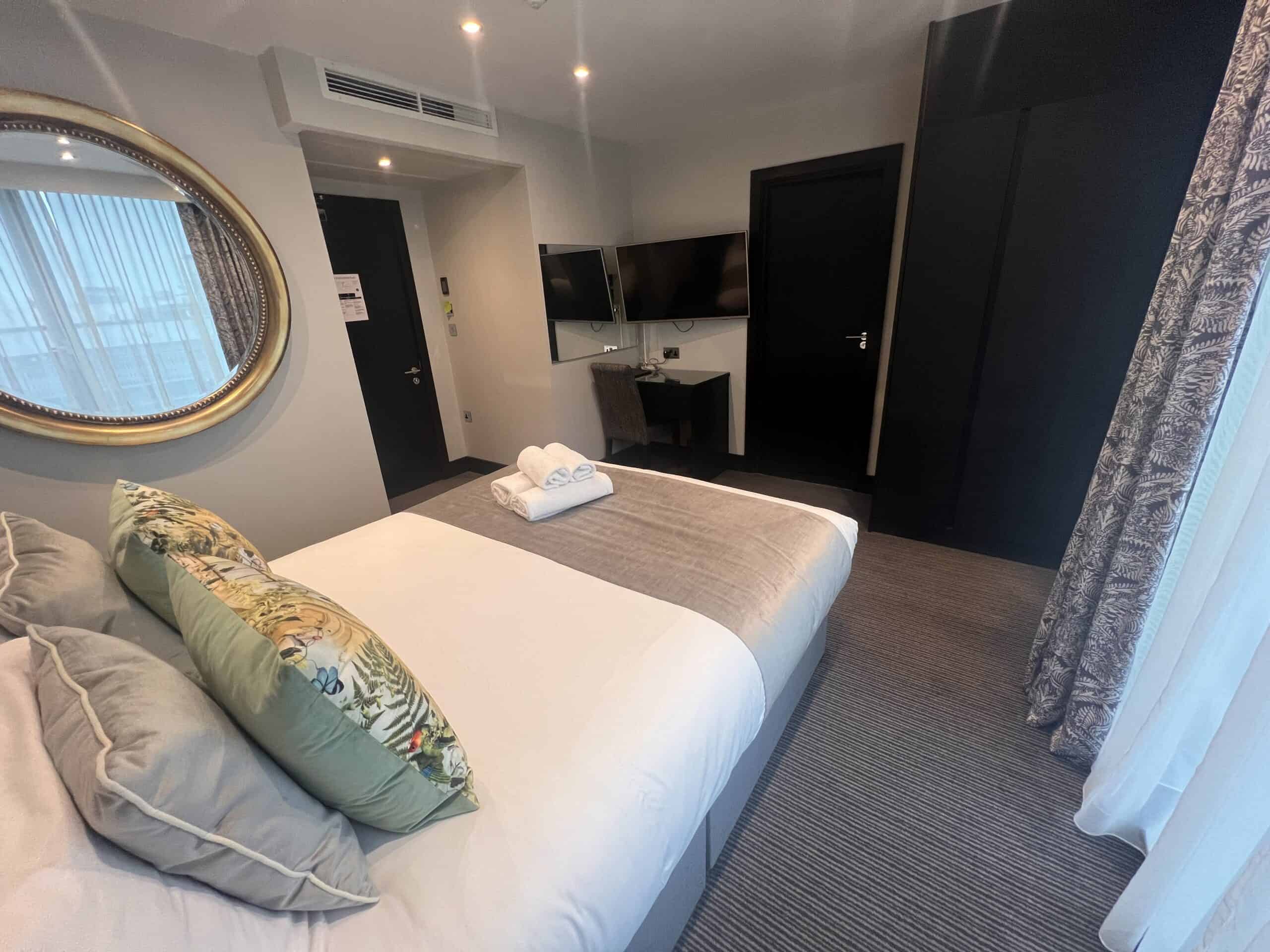 A modern hotel room featuring a neatly made bed with decorative pillows and a folded set of towels. A large round mirror hangs on the wall beside the bed. The room also has a flat-screen TV, a desk, and a window with patterned curtains.