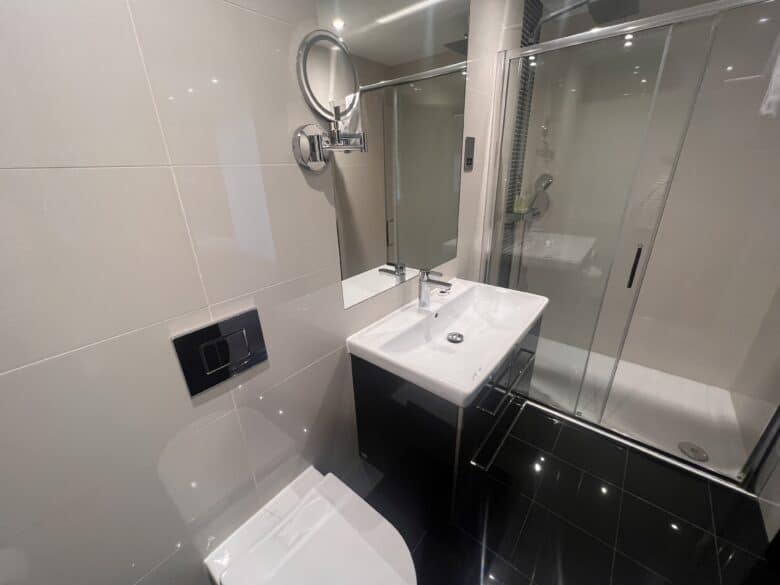 A modern bathroom featuring a white toilet, a rectangular sink with cabinets underneath, a circular wall-mounted mirror, and a walk-in shower with a glass door. The walls are light gray, and the floor is tiled in glossy black.