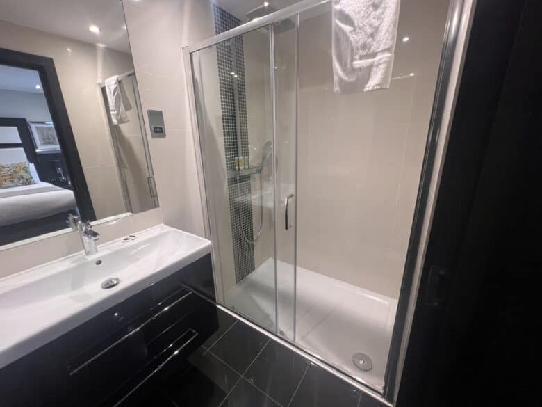 Modern bathroom with a glass-enclosed walk-in shower featuring a towel on the door. Adjacent, a sleek black vanity with a white sink and mirrored cabinet. The room extends to a visible bedroom with a bed and decorative cushions. Walls and floor have a clean finish.