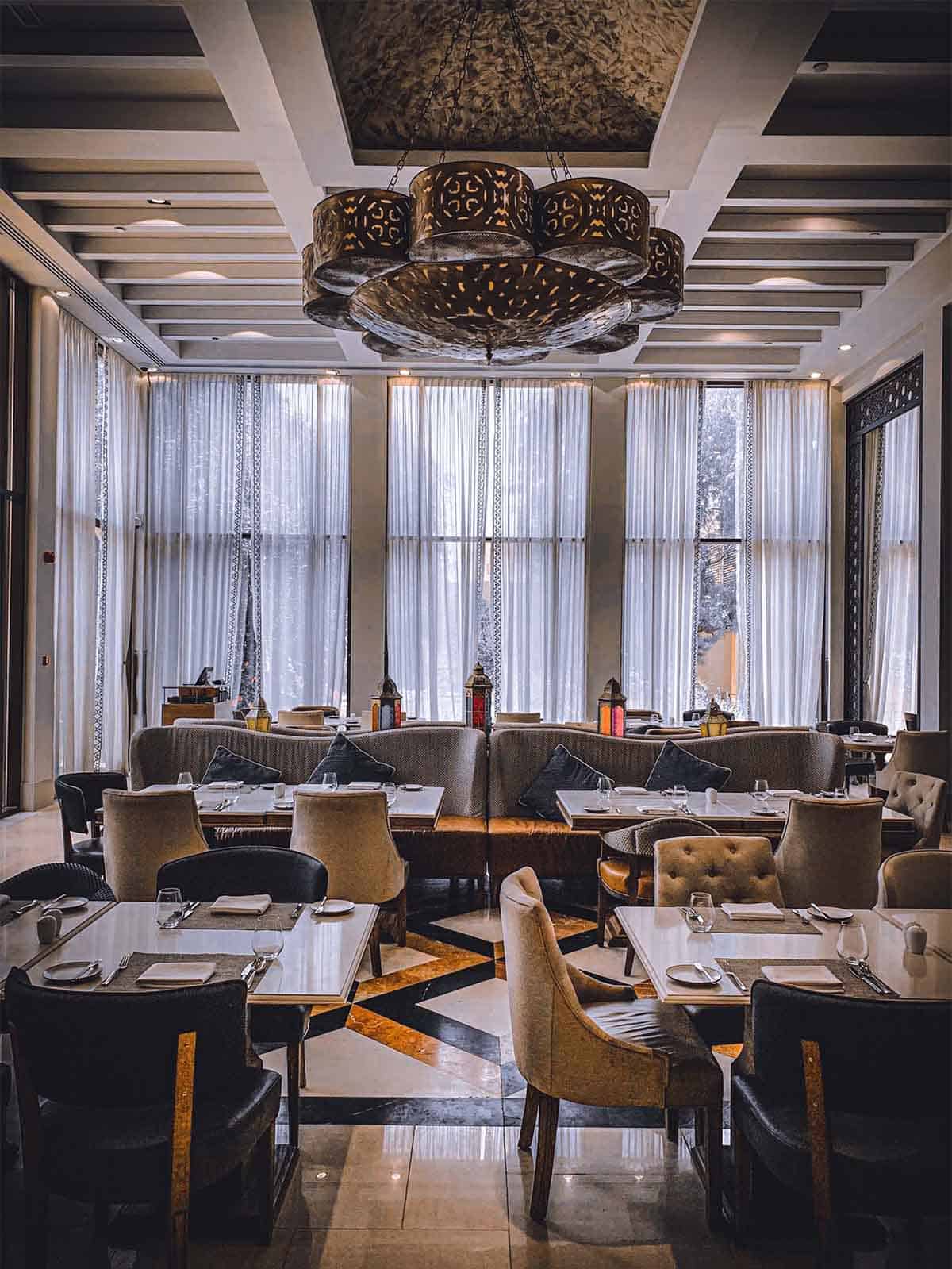 A sophisticated, modern restaurant interior within one of the finest hotels in London features several elegantly set tables and plush chairs. Large windows with sheer curtains allow natural light to stream in, illuminating the ornate ceiling design and a large decorative chandelier above.