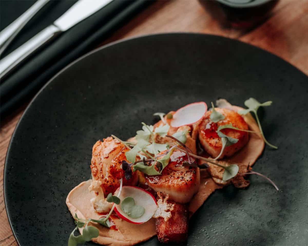 A black plate holds a gourmet dish featuring seared scallops on a puree, garnished with microgreens and thinly sliced radishes. Silverware and a rolled black napkin are in the background. The presentation is elegant and artistic, reminiscent of the fine dining experiences offered at top hotels in London.