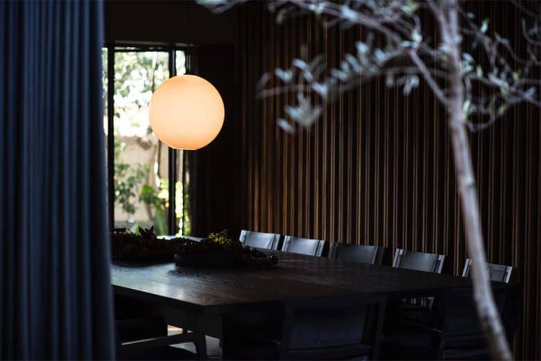 Dimly lit dining room with a large dark wooden table surrounded by dark chairs. A glowing spherical pendant light hangs above the table. Large windows in the background let in natural light, partially illuminating the space and revealing plants outside—a perfect setting reminiscent of boutique hotels in London.