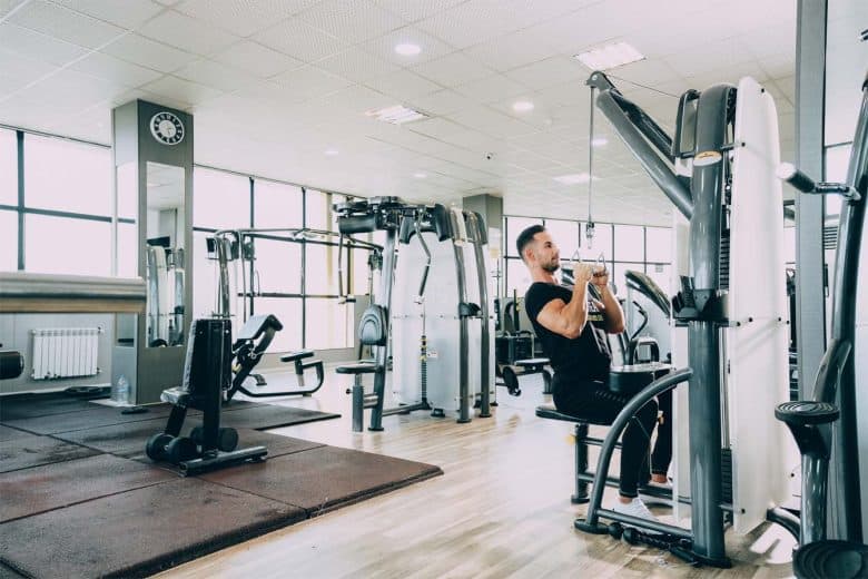 A person is seated on a machine in a spacious gym, performing a lat pulldown exercise. The gym, reminiscent of those found in upscale hotels in London, has various fitness equipment, large windows, and mirrored walls, with bright overhead lighting illuminating the space.