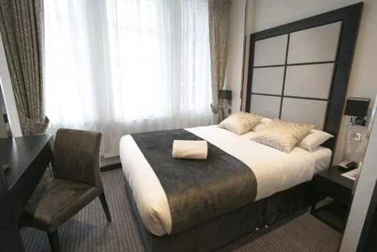 A hotel room in London with a neatly made double bed featuring two patterned pillows and a folded white towel. The room has dark wooden furniture, including a chair and a small table, and is softly lit by natural light from large windows with patterned curtains.