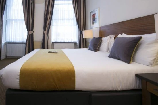 A neatly made bed with white linens, a yellow blanket, and gray pillows is in the center of a hotel room in one of the charming hotels in London. Natural light filters through two windows with dark curtains. A framed picture hangs on the wall above a wooden headboard, and a remote control lies on the bed.