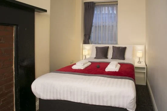 A cozy bedroom with a neatly made double bed, typical of charming hotels in London. The bed features a white duvet, red runner, and gray accent pillows. Towels rest at the foot, with a remote between them. Two bedside tables with lamps are on each side, and draped curtains frame the window above the headboard.