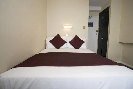 A cozy bedroom with a neatly made double bed featuring white linens, two maroon accent pillows, and a matching maroon throw blanket. A wall-mounted reading light is above the bed, reminiscent of elegant hotels in London. A closed closet door is on the right side of the room.