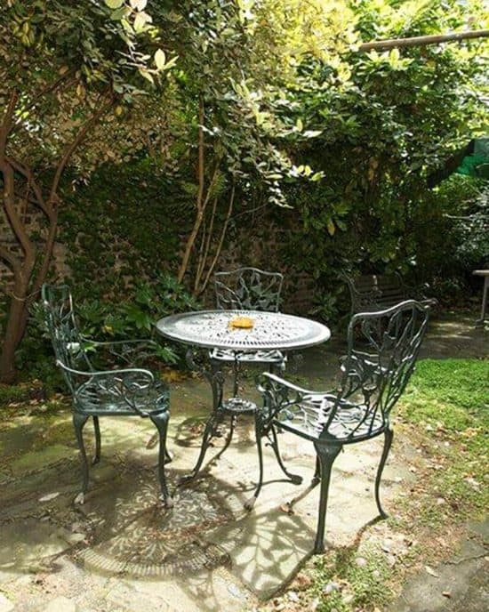 An ornate, wrought iron table and four matching chairs are set on a stone patio in a lush, leafy garden. Sunlight filters through the trees, casting intricate shadows on the ground. A small object, possibly a candle, sits at the center of the table—a scene reminiscent of tranquil spots found at some charming hotels in London.