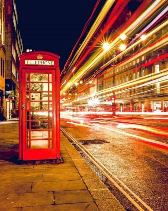 A classic red British telephone booth stands on a nighttime city street, with light trails from passing vehicles creating vibrant streaks of color. Nearby hotels in London line the street under the dark sky, emphasizing the dynamic, bustling urban environment.