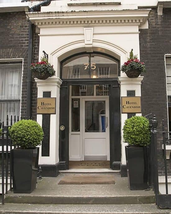 Entrance to Hotel Cavendish with double white doors framed by black trim and a white arch. The number 75 is displayed above the doors. Two potted plants are positioned beside the entrance, along with hanging flower baskets. As one of the charming hotels in London, its name is elegantly displayed on placards.