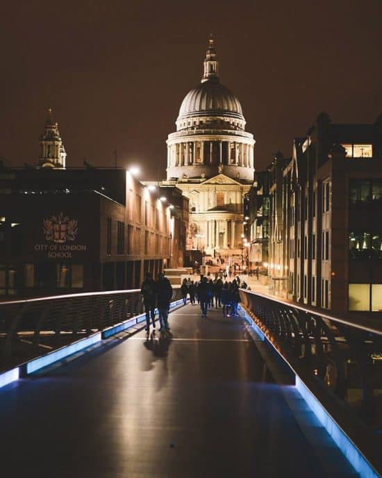 A nighttime view of St. Paul's Cathedral in London, beautifully illuminated. People walk along a modern, lit bridge leading towards the historic building, creating a striking contrast between old and new architecture. The City of London School is visible on the left, with nearby hotels in London completing the scenery.