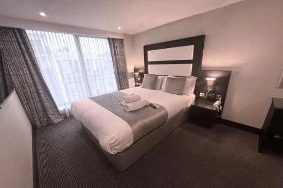 A cozy hotel room with a large bed topped with neatly folded towels and remote control. The room features two bedside tables with lamps, a floor-to-ceiling window with sheer curtains, and a dark wooden headboard with white panels. The overall decor is modern and stylish, perfect for those seeking hotels in London.