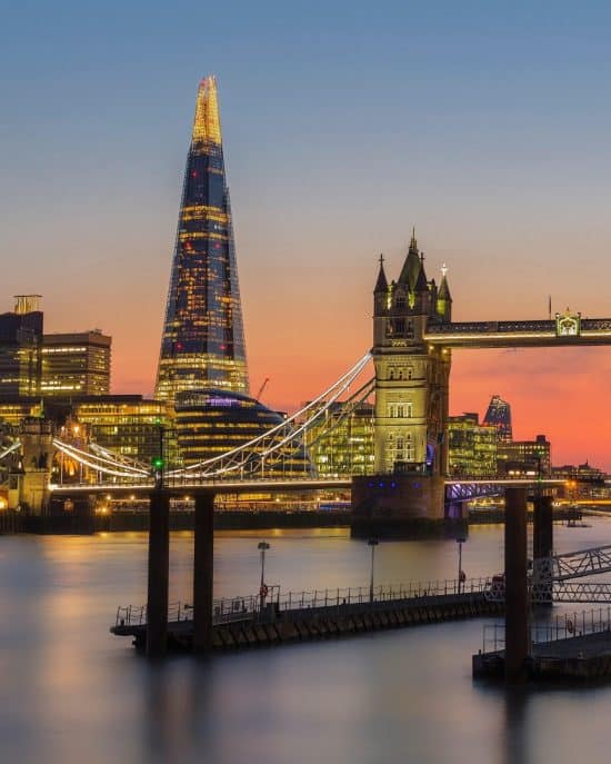 A vibrant sunset over London, featuring the illuminated Shard skyscraper and Tower Bridge. The bridge's gothic towers and cables are visible over the calm waters of the Thames, with the city's skyline forming an impressive backdrop, promising a memorable evening for guests at nearby hotels in London.