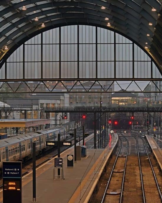 A train station platform under a large arched roof, with a view of tracks curving away into the distance. A train is at the platform and digital display screens show departure times. The background shows city buildings and the sky is overcast, with signs pointing to nearby hotels in London.